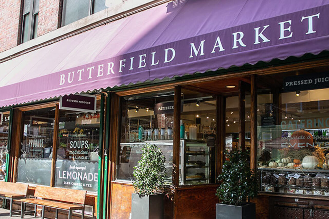 Purple awning lettered 'Butterfield Market' over a wood-framed grocery storefront, with soup, salad and lemonade decals in the windows and wooden benches on the sidewalk.