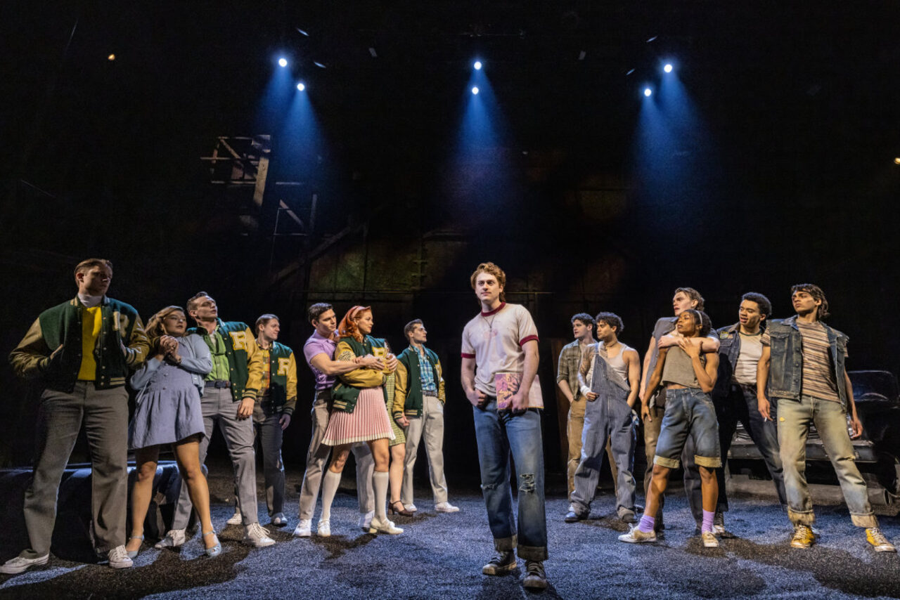 The full cast of a Broadway musical on a dark stage under blue spotlights — Greasers in denim and leather facing Socs in letterman jackets, the lead actor center stage holding a paperback novel.