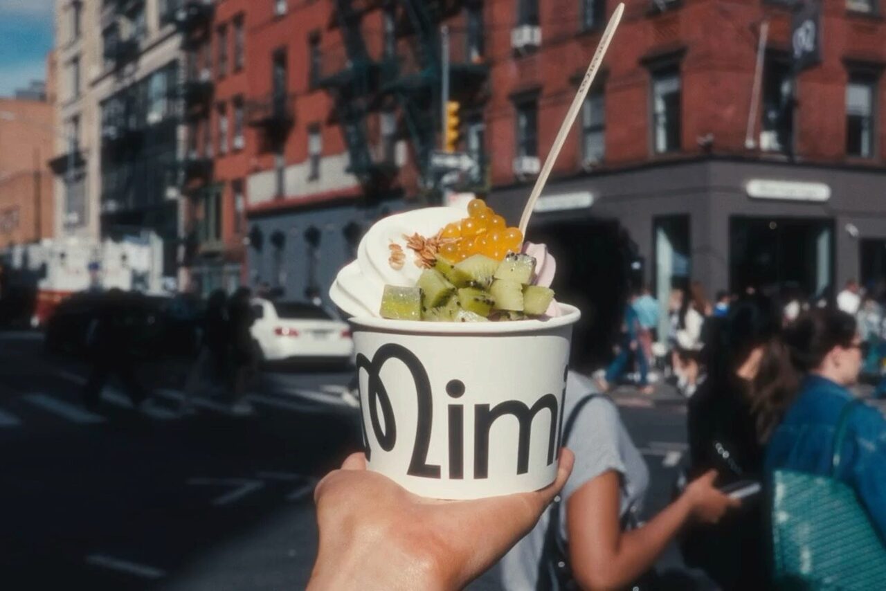 A paper cup of frozen yogurt topped with kiwi, popping boba and granola — the Mimi's wordmark printed on the cup — held up on a sunny Manhattan street corner.